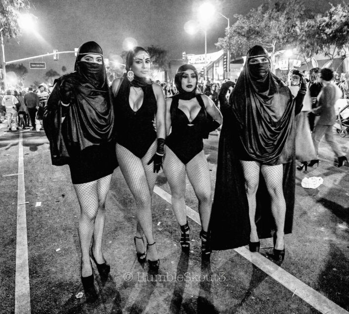 Four Arab/ Muslim females in fishnet stockings, heels, bodysuits, short skirts topped off with either a: Turban, Snood Hijab, the Hijab or niqab or headscarf at the Halloween Carnaval, West Hollywood. Open Rebellion (Los Angeles Street Photography) (black and white)