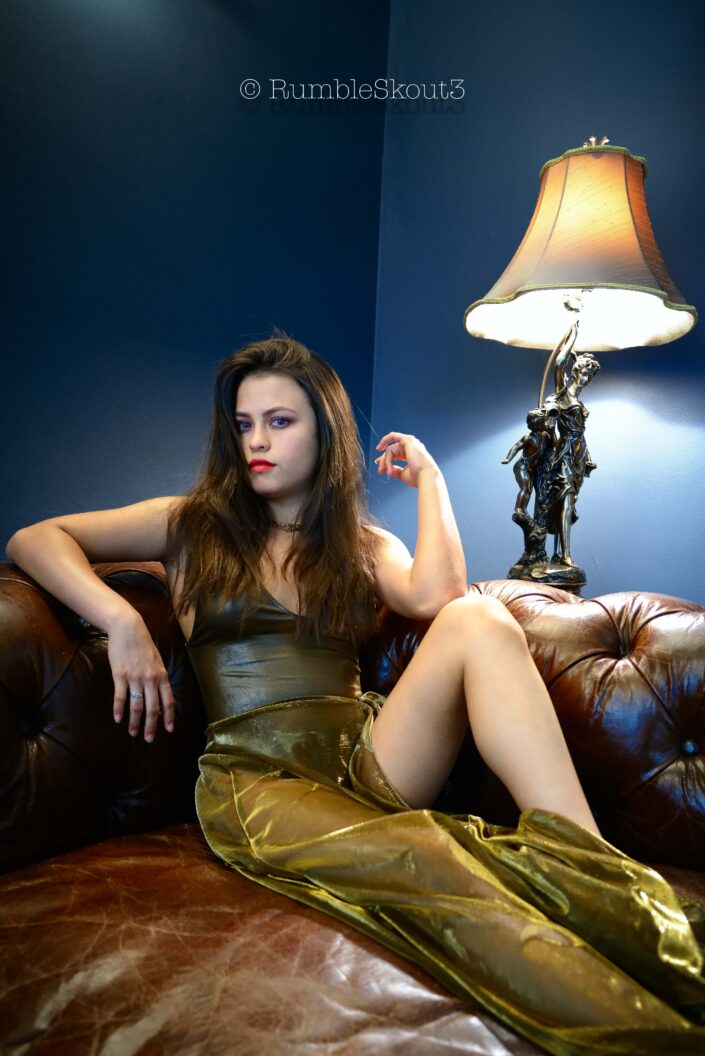 An American Élégance Bella in an Art Déco Azul Salon, with amber eyes, in a bodysuit sheer dress, on a leather chesterfield sofa sitting for an American woman portrait in West Hollywood, contemplating Mars.