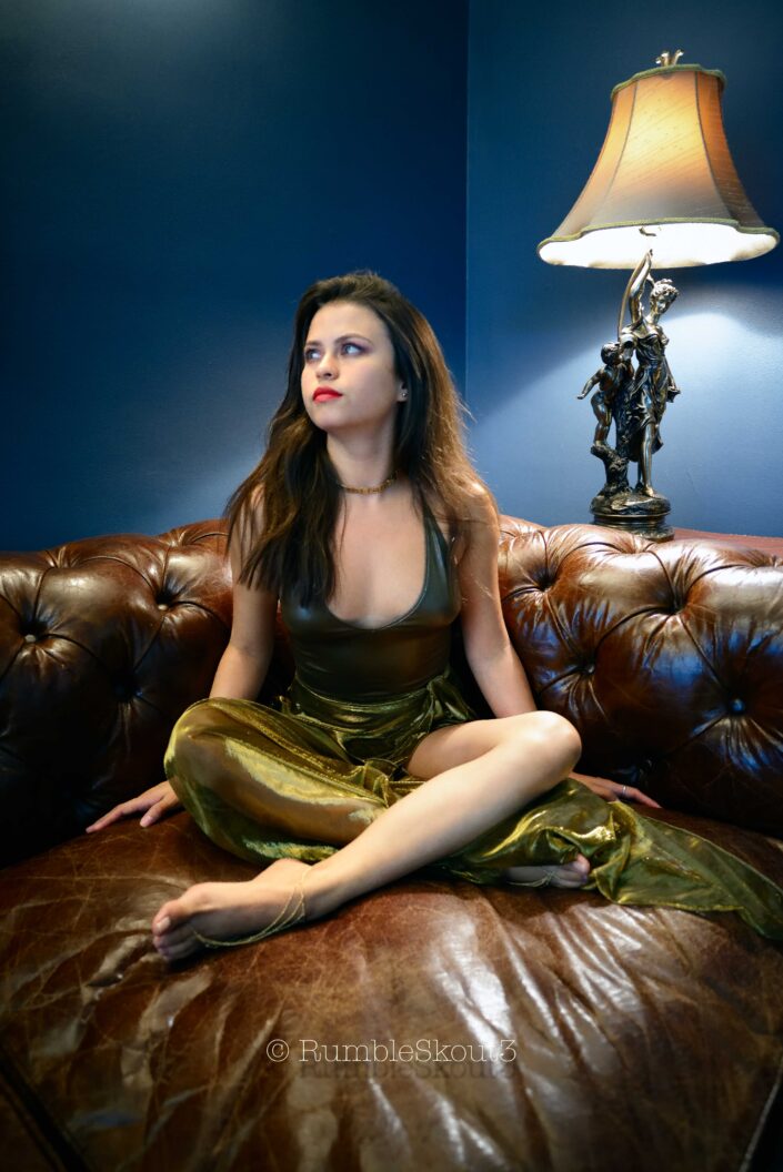 An American Élégance Bella in an Art Déco Azul Salon in a bodysuit sheer dress, on a leather chesterfield sofa sitting for an American woman portrait in West Hollywood, contemplating Mars.