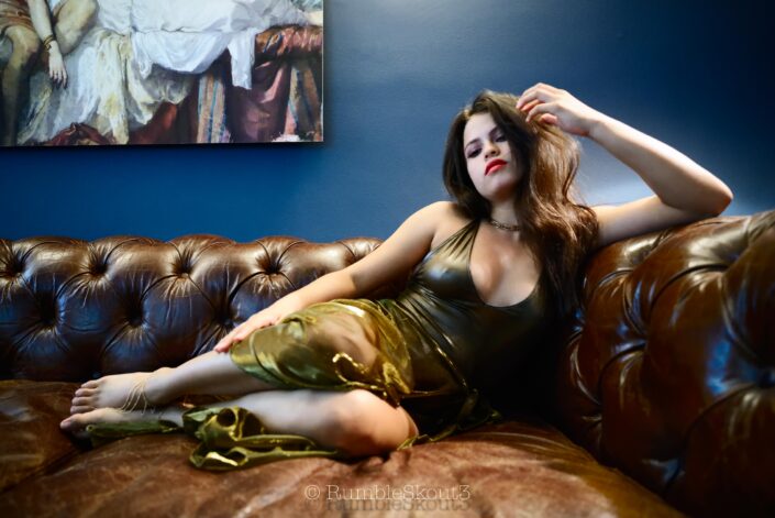 An American Élégance Bella in an Art Déco Azul Salon in a bodysuit sheer dress, on a leather chesterfield sofa sitting for an American woman portrait in West Hollywood, contemplating Mars.